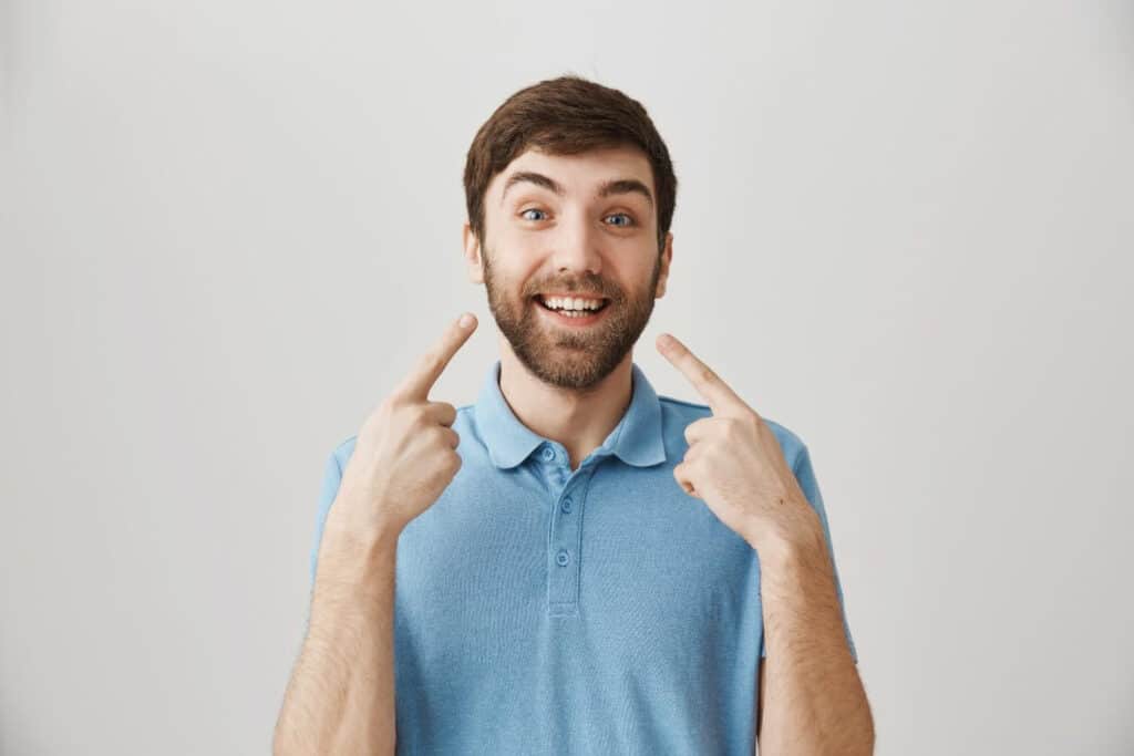 Uomo sorridente in camicia blu che si punta i denti, mostrando felicità e fiducia su uno sfondo semplice.