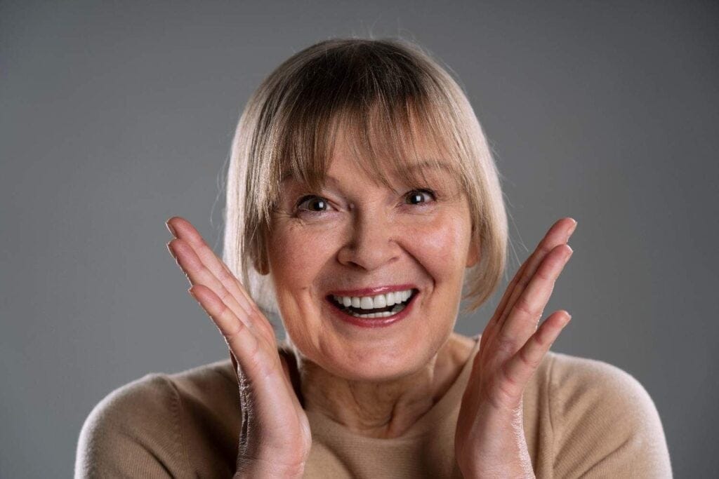 Elderly woman smiling with joy, hands raised excitedly near face against gray background.
