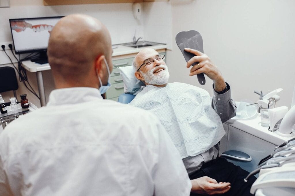Burrë i moshuar duke i buzëqeshur dentistit, duke kontrolluar dhëmbët me pasqyrë dore në zyrën dentare.