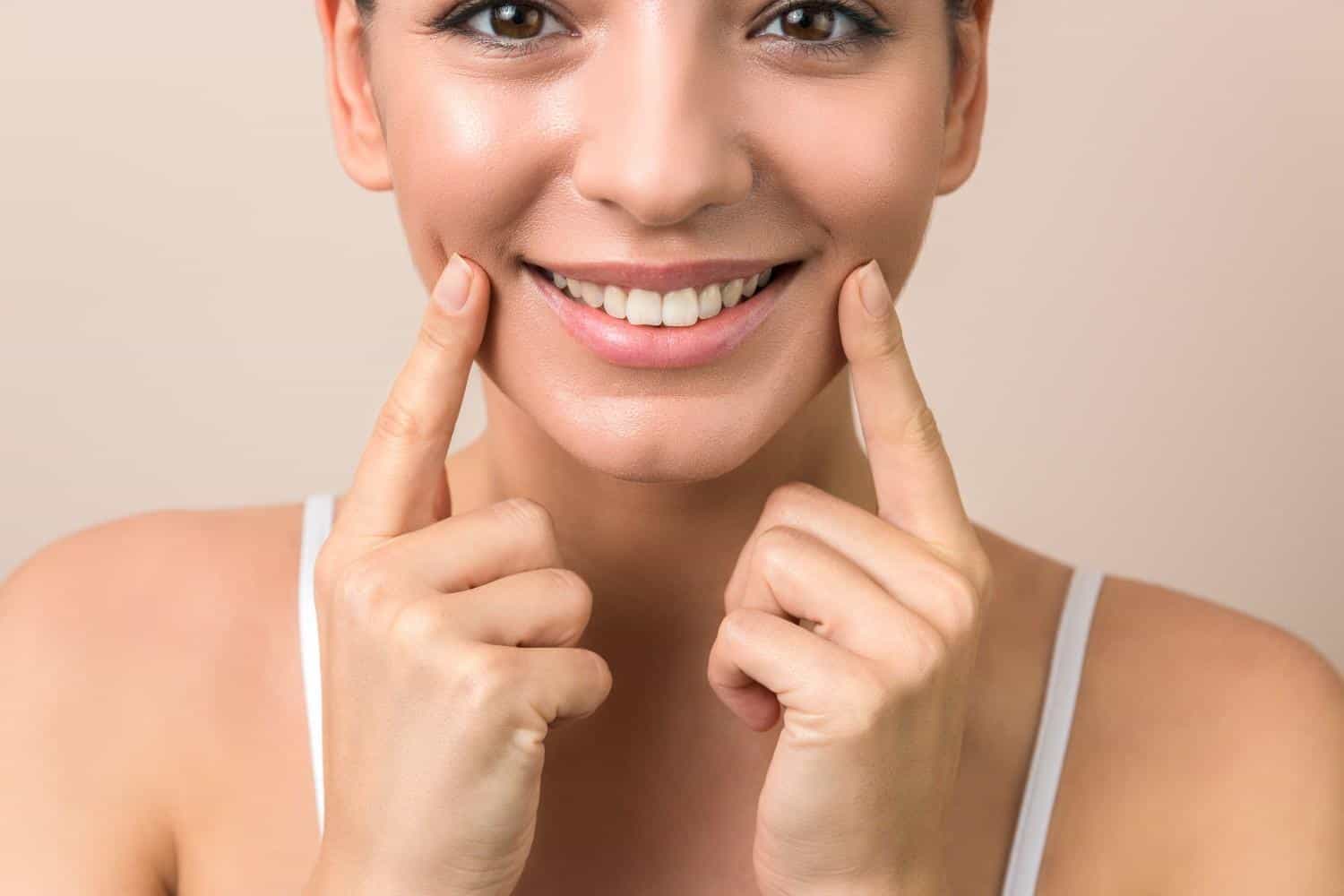 Femme souriante se touchant les joues, gros plan sur les dents et la peau, mettant en avant la santé dentaire et les soins de la peau.
