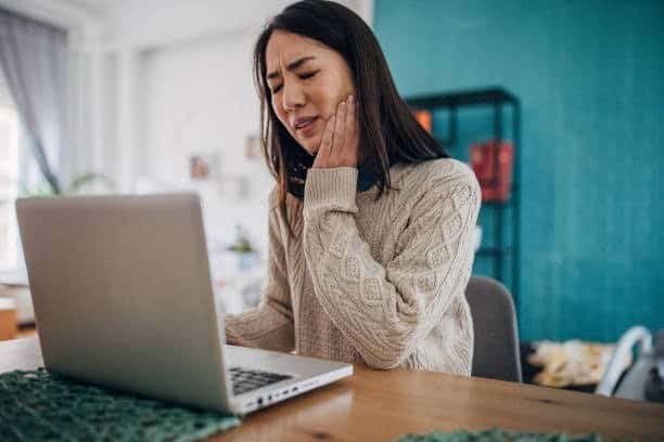 Woman experiencing toothache while using laptop at home.
