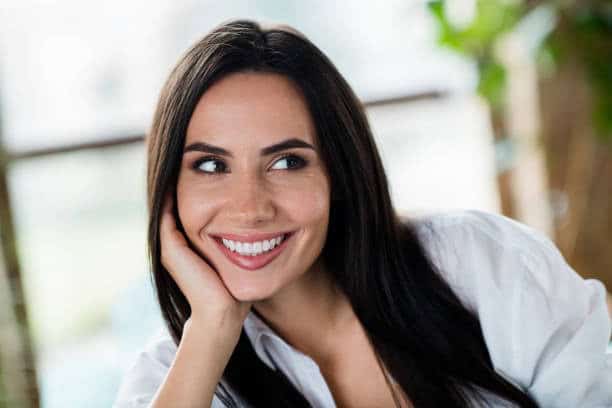 Smiling woman with long dark hair in white shirt, looking to the side, sitting indoors with natural light and plants in background.