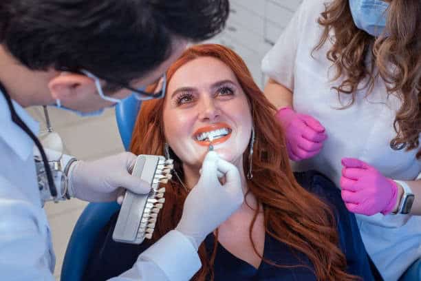 Woman in dental chair receiving teeth whitening treatment from dentist and assistant in clinic setting.