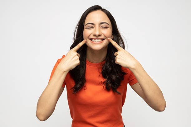 Smiling woman in an orange shirt pointing at her teeth, showcasing a bright, cheerful expression against a light background.