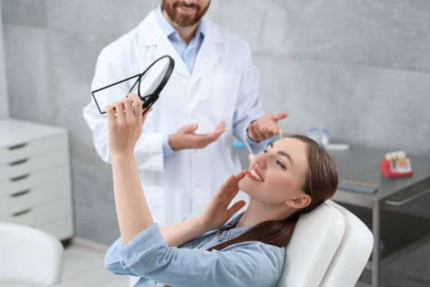 Woman admiring smile in mirror at dental clinic with dentist. Happy patient after dental check-up.