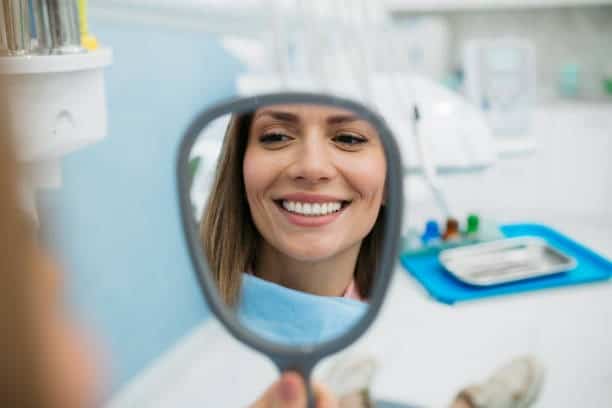 Woman smiling at dental office, holding mirror to admire teeth. Happy with dental care results.