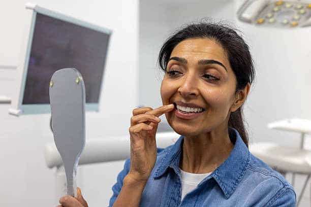 Woman at dentist's office smiling and examining her teeth with a handheld mirror.